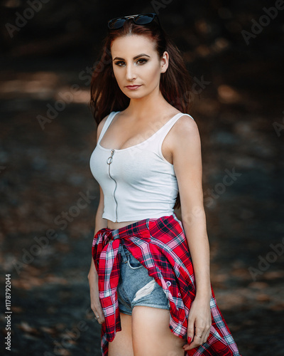 woman with long brown hair. She is wearing a white tank top with a zipper on the front and denim shorts. She has a red plaid shirt tied around her waist. She has sunglasses on her head, pulled up by h photo