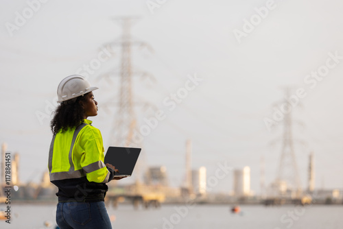 A female engineer in a high-visibility jacket and helmet uses a laptop and smartphone near a power plant and transmission towers, illustrating modern technology, communication, and industrial work