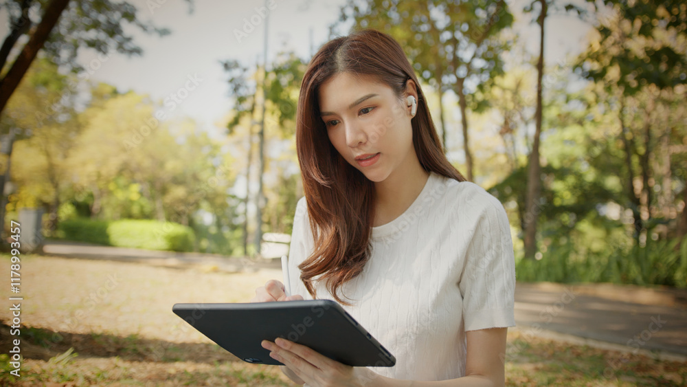 Obraz premium Asian Woman Working outdoors in the park. Professional on a Video Call, exploring Hybrid Work Opportunities in a Peaceful, Virtual Meeting and Online Learning. Hybrid work lifestyle concept by tablet