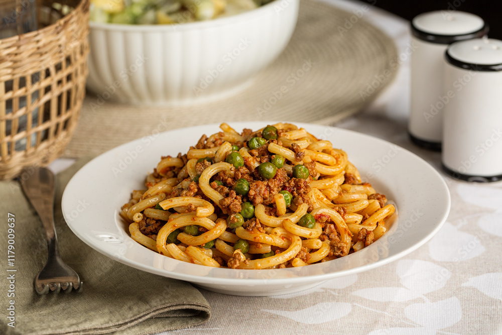 Plate with italian pasta gramigna with meat and tomato ragu sauce and green peas. Table served for lunch, salad and bottle of water on background. Plate with italian pasta gramigna with meat and tomato ragu sauce and green peas. Table served for lunch, salad and bottle of water on background.