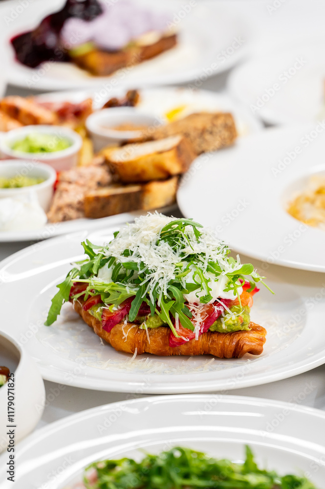 A freshly baked croissant topped with avocado, arugula, and shaved cheese, served on a white plate. In the background, a variety of colorful dishes create an appealing brunch setting. A freshly baked croissant topped with avocado, arugula, and shaved cheese, served on a white plate. In the background, a variety of colorful dishes create an appealing brunch setting.