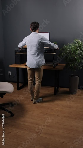 Workstation setup, Standing desk, Desk ergonomic. In modern office with wooden flooring and ergonomic chair, young man in floral hoodie is adjusting standing desk, Vertical video.