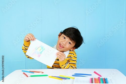 A cheerful boy proudly displays his colorful drawing on white paper, sitting at a table filled with vibrant crayons. Wearing a yellow striped shirt, he beams with creativity against a blue background. photo