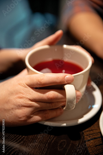 A pair of hands gently holding a white ceramic cup filled with red tea, placed on a wooden table. The warm, cozy moment reflects comfort and relaxation in a minimalist setting.