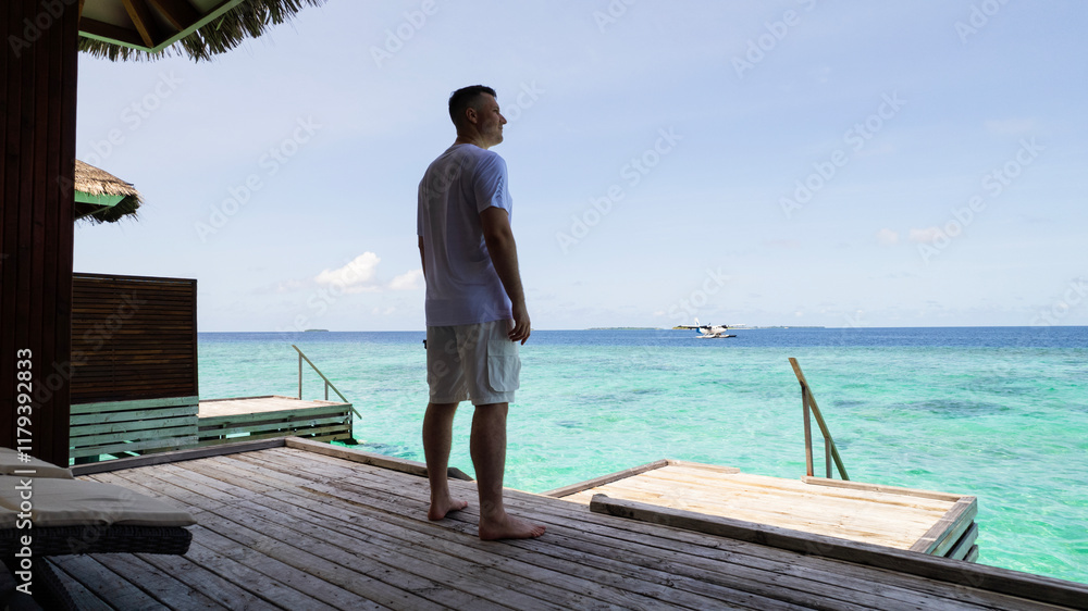 A guy in white clothes admires the turquoise ocean. Vacation in the Maldives. He looks into the future and enjoys it. Small waves. Descent into the water. Wooden villa. Bungalows on the water