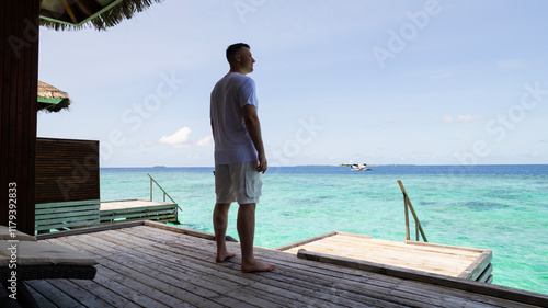 Obraz na płótnie A guy in white clothes admires the turquoise ocean. Vacation in the Maldives. He looks into the future and enjoys it. Small waves. Descent into the water. Wooden villa. Bungalows on the water w salonie