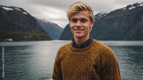 Blond Norwegian man enjoys a tranquil moment by the serene fjord surrounded by majestic mountains under a cloudy sky