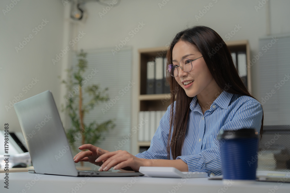 Young Asian businesswoman wearing glasses, smiling while working on a laptop and calculator in a modern office. Engaging with the screen, she exudes professionalism and confidence Young Asian businesswoman wearing glasses, smiling while working on a laptop and calculator in a modern office. Engaging with the screen, she exudes professionalism and confidence
