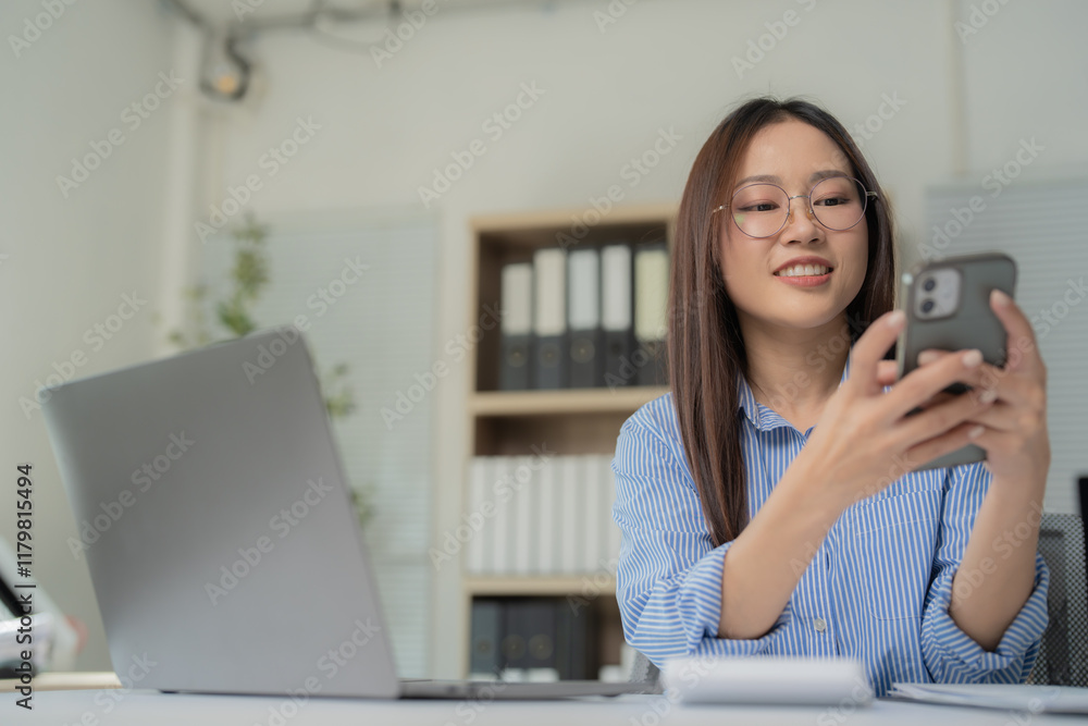 Young Asian businesswoman wearing glasses, smiling while using a smartphone and working on a laptop at a modern office desk, managing her business and checking social media Young Asian businesswoman wearing glasses, smiling while using a smartphone and working on a laptop at a modern office desk, managing her business and checking social media