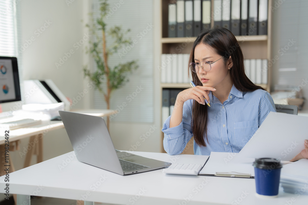 Focused Asian businesswoman reviewing documents and analyzing data with a laptop and calculator at her office, thoughtfully planning online projects and strategies Focused Asian businesswoman reviewing documents and analyzing data with a laptop and calculator at her office, thoughtfully planning online projects and strategies