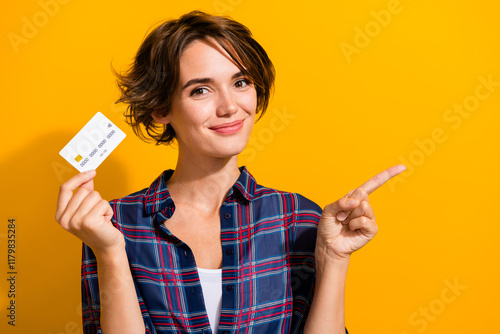 Photo of positive woman dressed checkered shirt hold credit card indicating at discount empty space isolated on yellow color background photo