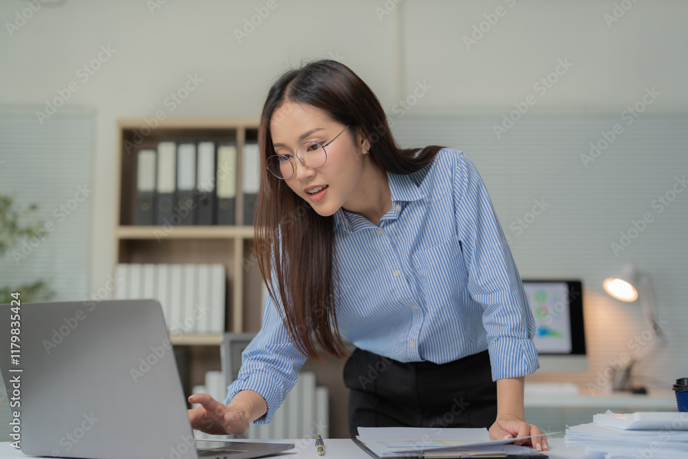 Young asian businesswoman wearing glasses standing at her desk working using laptop computer, focused business woman working with documents and typing on laptop in office Young asian businesswoman wearing glasses standing at her desk working using laptop computer, focused business woman working with documents and typing on laptop in office
