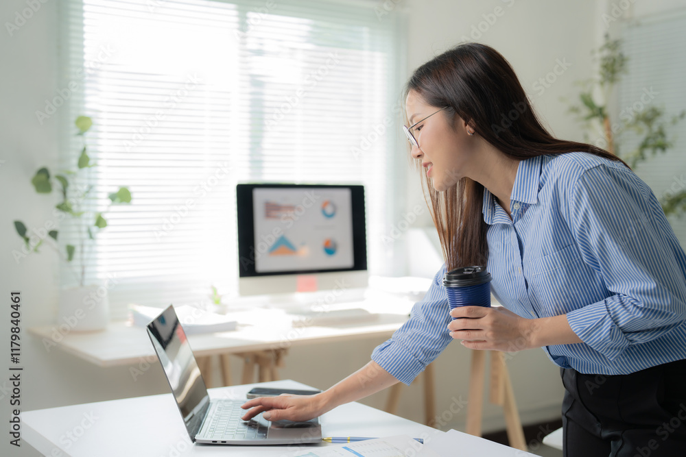 Young asian businesswoman holding a coffee and working on laptop in modern office with desktop computer showing graphs on the screen in the background Young asian businesswoman holding a coffee and working on laptop in modern office with desktop computer showing graphs on the screen in the background