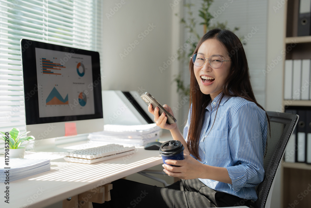 Young Asian businesswoman working with computer and smartphone while drinking coffee and reading good news on her mobile phone, feeling happy and cheerful, sitting at desk in modern office Young Asian businesswoman working with computer and smartphone while drinking coffee and reading good news on her mobile phone, feeling happy and cheerful, sitting at desk in modern office