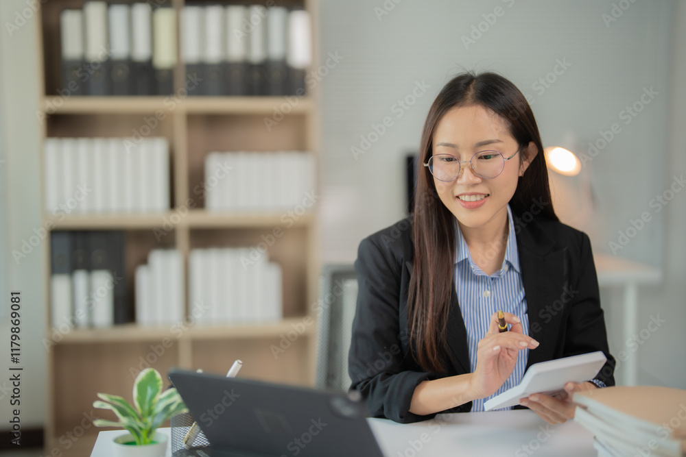 Young professional using calculator and tablet while working on a project in a modern office with bookshelves in the background, showcasing a focused and productive work environment Young professional using calculator and tablet while working on a project in a modern office with bookshelves in the background, showcasing a focused and productive work environment