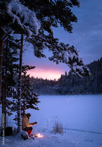 Man sitting and drinking coffee near a campfire in a winter landscape with the glowing sky after the sunset behind