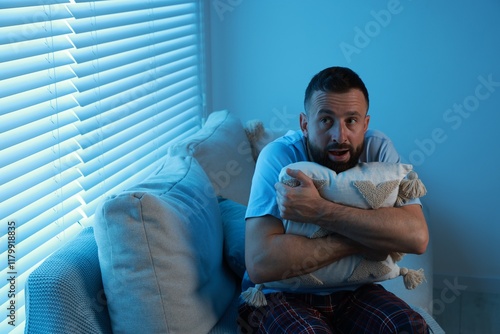 Fear of darkness. Scared man hugging pillow on couch in dark room, space for text photo