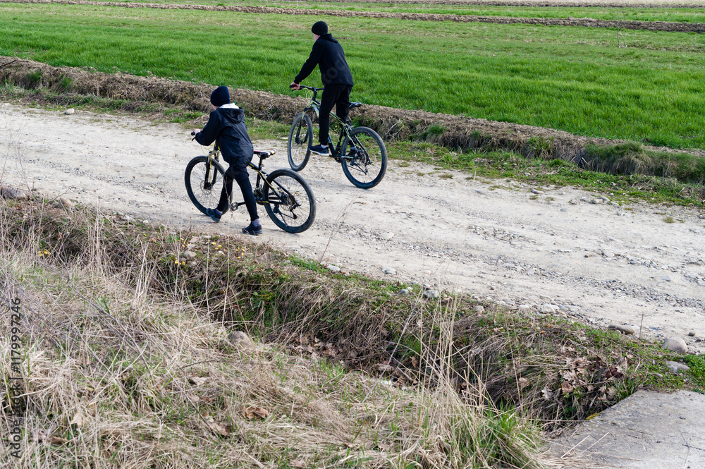 Two figures on bicycles traverse a dirt path bordering a verdant field. The sunlit scene evokes tranquility and outdoor activity. Two figures on bicycles traverse a dirt path bordering a verdant field. The sunlit scene evokes tranquility and outdoor activity.