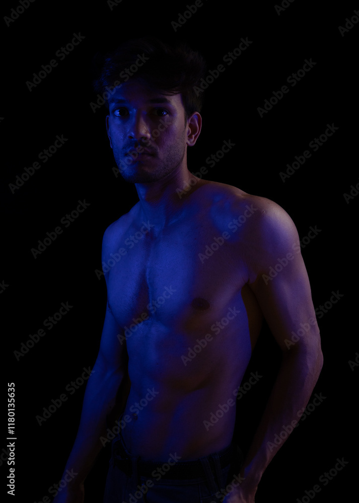 Fototapeta premium Junger Mann aus Indien steht mit freiem Oberkörper in buntem Licht und posiert