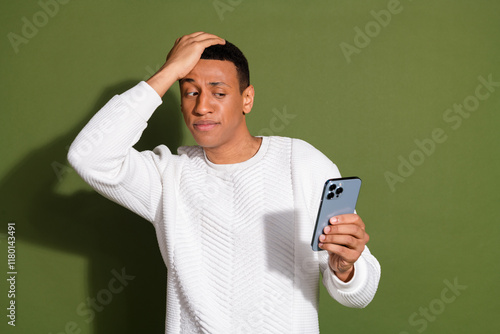 Tablou pe pânză Young man in a sweater looks puzzled at smartphone against khaki backdrop, showc
