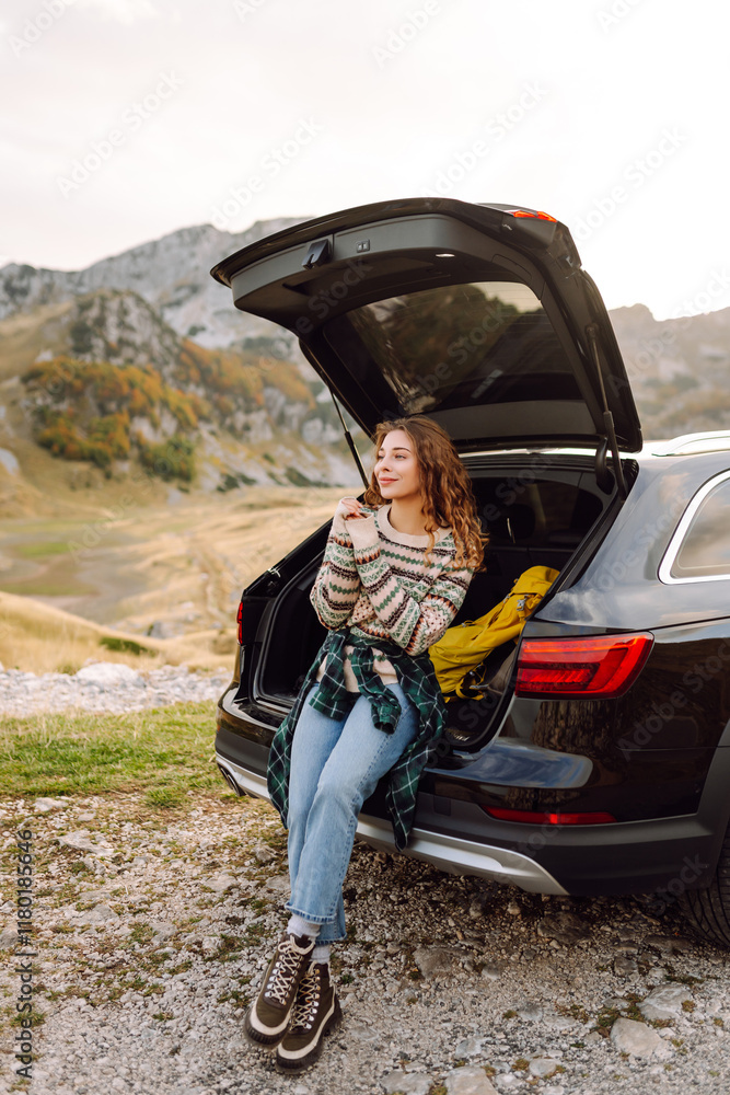 Naklejka premium Happy woman sits in the trunk of her car, parked on the side of a winding road that cuts through the rocky terrain. Lifestyle, travel, tourism, nature, active life