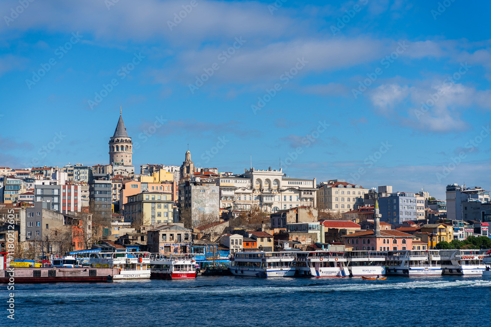 Fototapeta premium The Galata Tower rises high above the rooftops of Istanbul with tourist boats on the banks of the Bosphorus in the foreground