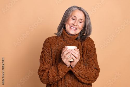 Charming senior woman holding a mug and smiling joyfully against a beige background in casual brown knitwear.