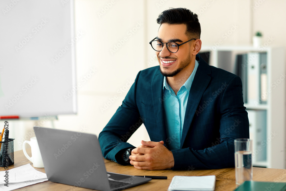 Confident young Arab entrepreneur in formal suit using laptop computer at his desk, having virtual meeting at office. Millennial businessman using pc for online job, communicating remotely Confident young Arab entrepreneur in formal suit using laptop computer at his desk, having virtual meeting at office. Millennial businessman using pc for online job, communicating remotely