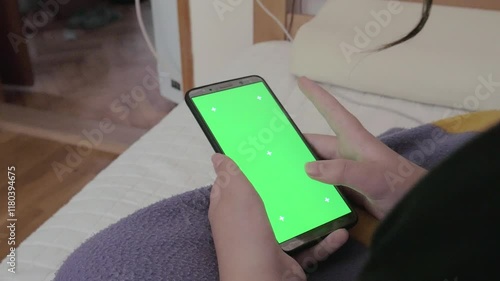 A close-up shot of a girl's hand holding a smartphone and scrolling through the screen. The phone screen is a green screen with tracking markers. Handheld shot, over the shoulder