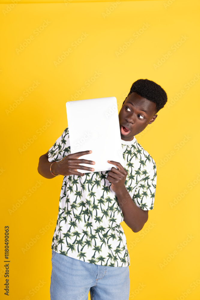 Surprised young man hiding behind laptop showing blank screen on yellow background Surprised young man hiding behind laptop showing blank screen on yellow background