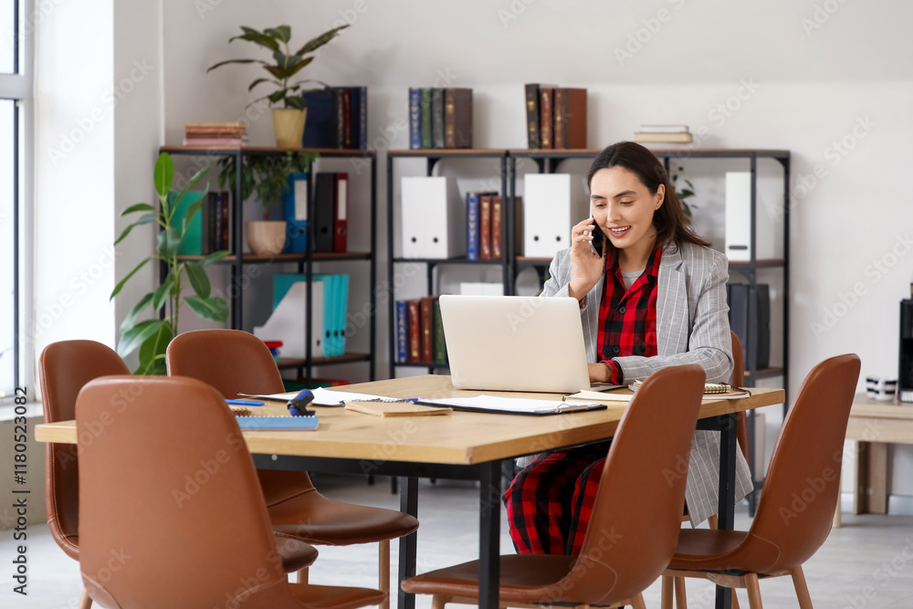 Young businesswoman talking by mobile phone at table in office. National Wear Your Pajamas to Work Day Young businesswoman talking by mobile phone at table in office. National Wear Your Pajamas to Work Day