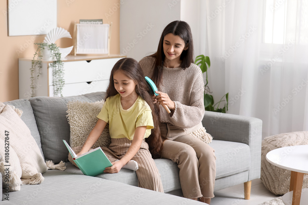 Young mother brushing hair of her little daughter who reading book while sitting on sofa at home Young mother brushing hair of her little daughter who reading book while sitting on sofa at home