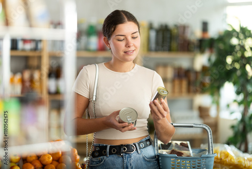 Young woman is holding two tin cans of canned food in hands, choosing pate for sandwiches. She carefully studies information for consumer, thinks about buying photo