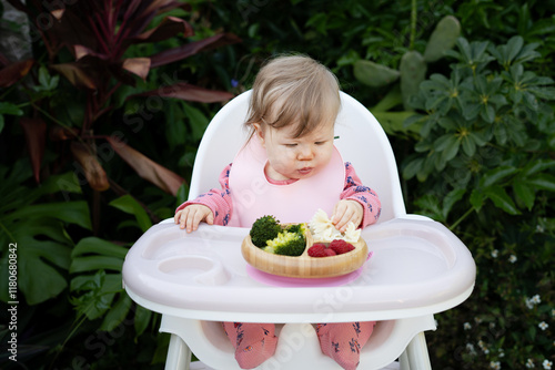 Cute Caucasian baby girl sitting in a high chair and eating healthy food. Baby-led weaning concept. Baby eating finger foods in a tropical background, with copy space. Baby eating pasta photo