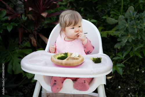 Cute Caucasian baby girl sitting in a high chair and eating healthy food. Baby led weaning concept. Baby eating finger foods in a tropical background, with copy space. Baby eating raspberry photo