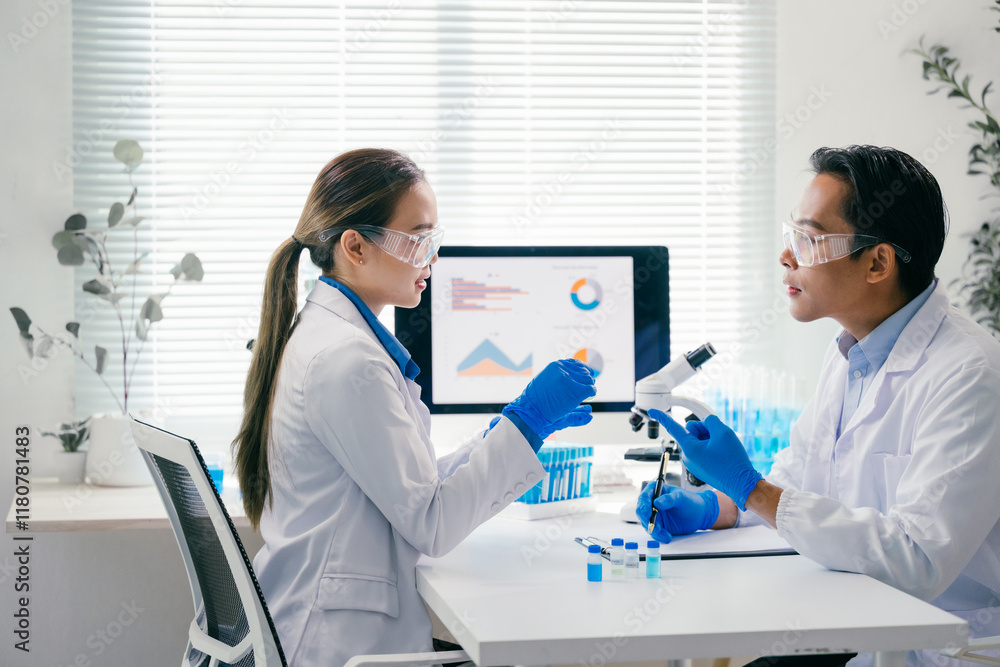 Two scientists in lab coats and safety glasses discussing research results at a table in a laboratory, surrounded by a microscope, test tubes, and a computer with charts Two scientists in lab coats and safety glasses discussing research results at a table in a laboratory, surrounded by a microscope, test tubes, and a computer with charts
