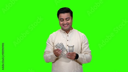 A young man counting cash notes in a happy mood. He has Indian currency. A green screen studio.
