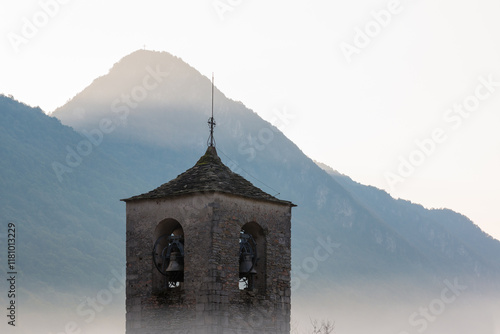 Romanesque bell tower emerges from the fog with mountains in the background and the sun about to rise. Bell tower (12th century ) of the Abbey of San Gemolo in Valganna, province of Varese, Italy  photo