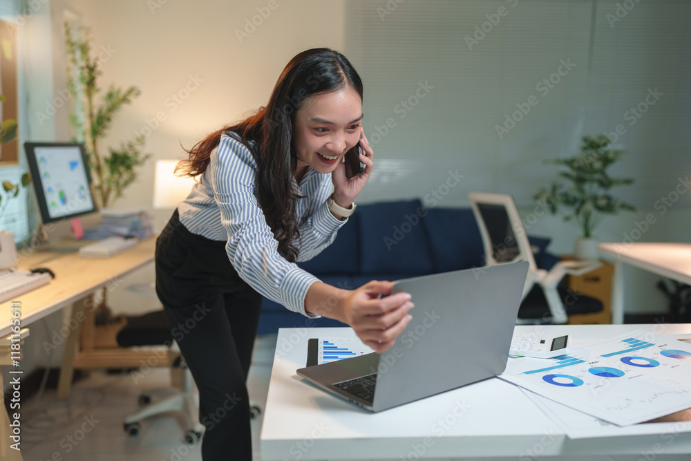 Obraz premium Young, happy asian businesswoman talking on a mobile phone while showcasing her laptop to a colleague, working late in the office at night, immersed in collaboration and technology