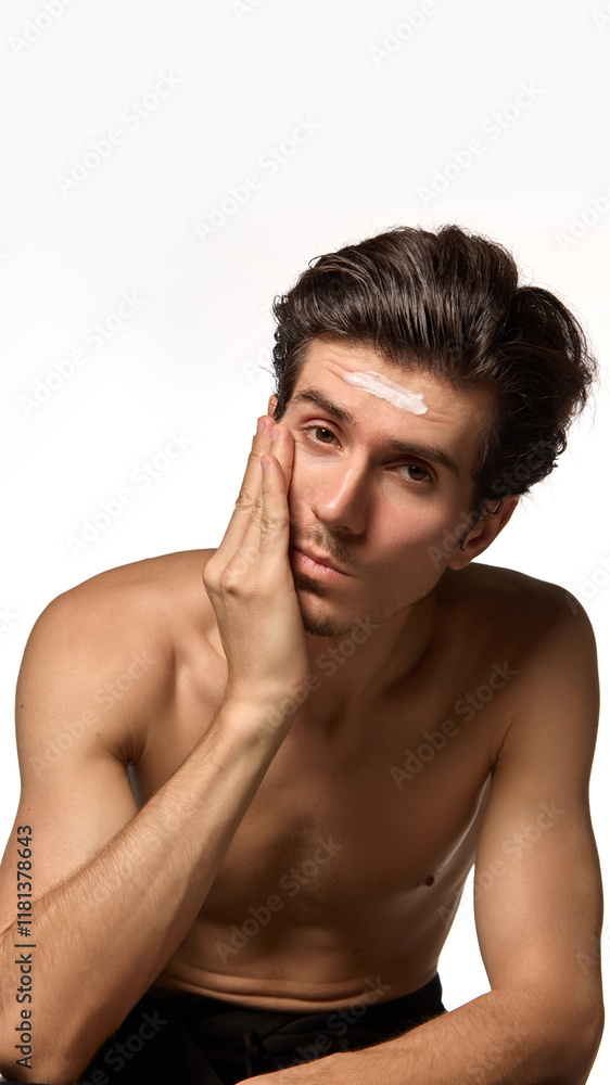 Obraz premium Young attractive man posing, sitting gently holding hand on cheek with cream on forehead against white background. Concept of male beauty and facial care, cosmetology, dermatology. Ad