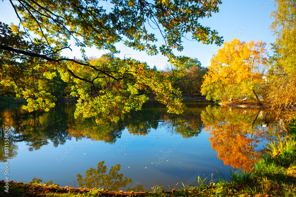 Serene lake surrounded by trees with autumn foliage, reflecting vibrant colors on the calm water surface. Clear sky and bright sunlight enhance beauty of scene, making it a picturesque autumn day Serene lake surrounded by trees with autumn foliage, reflecting vibrant colors on the calm water surface. Clear sky and bright sunlight enhance beauty of scene, making it a picturesque autumn day