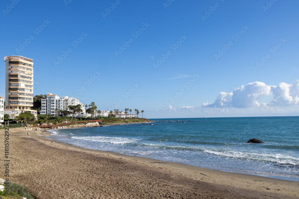 Photo of the beautiful beach front in the town of Sitio de Calahonda in Spain showing the ocean waves crashing in to the beach on a sunny day with a blue sky and a few small clouds Photo of the beautiful beach front in the town of Sitio de Calahonda in Spain showing the ocean waves crashing in to the beach on a sunny day with a blue sky and a few small clouds