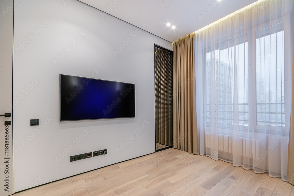 The living room in the apartment has a large window and an installed TV, unfurnished. .White room decor, beige laminate. The living room in the apartment has a large window and an installed TV, unfurnished. .White room decor, beige laminate.