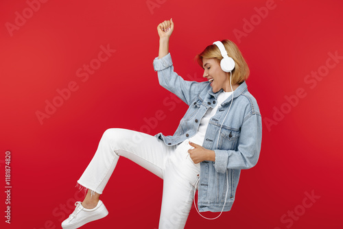 Stylish young blonde woman in casual white outfit listening music in headphones over red background.