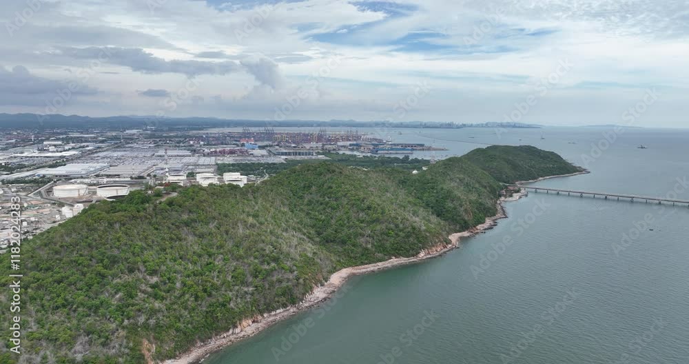 Drone flying over the ocean and green cliffs, revealing a petrochemical refinery under cloudy sky Drone flying over the ocean and green cliffs, revealing a petrochemical refinery under cloudy sky