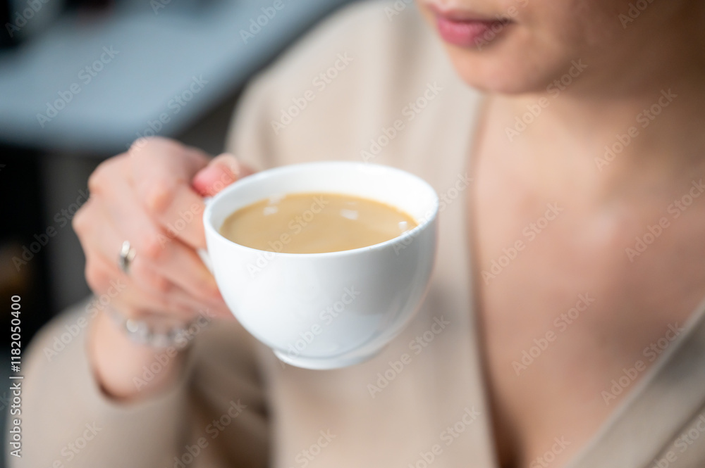 A close-up of a woman's hand holding a white cup of coffee. The focus is on the cup, with the woman's face slightly blurred in the background, creating a warm, cozy atmosphere. A close-up of a woman's hand holding a white cup of coffee. The focus is on the cup, with the woman's face slightly blurred in the background, creating a warm, cozy atmosphere.