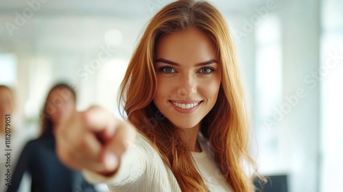 smiling young businesswoman with long auburn hair pointing at camera in modern office while diverse team members engage in background photo