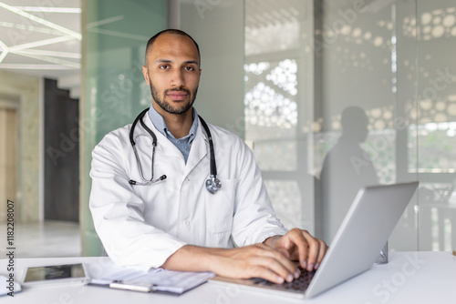 Portrait experienced doctor inside a clinic office at workplace, man is looking at the camera seriously and focused, working with a laptop, preparing an electronic report of the patient's treatment.