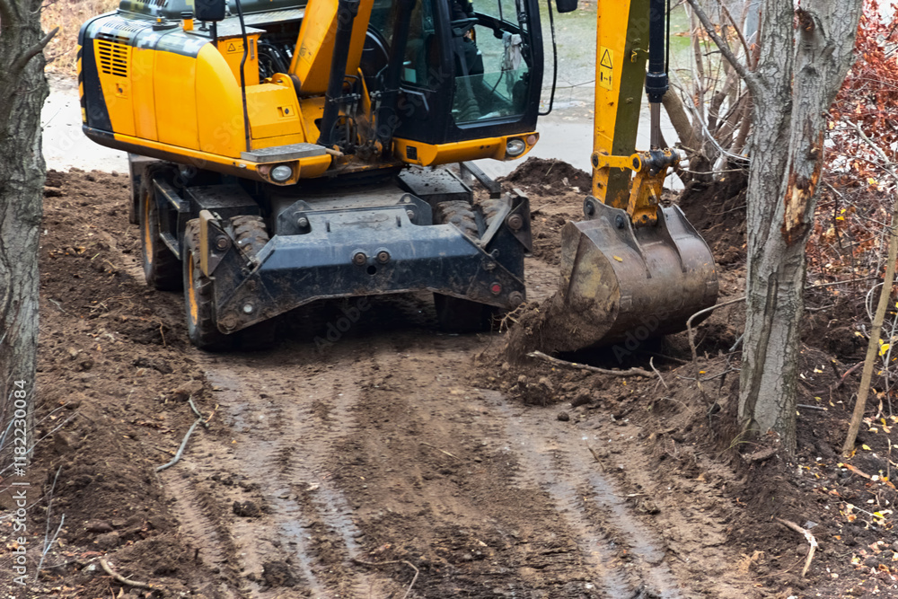An excavator digs the ground on a site with trees, demonstrating work in confined spaces and the need for careful treatment of the environment. An excavator digs the ground on a site with trees, demonstrating work in confined spaces and the need for careful treatment of the environment.