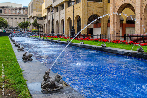 Fountain with jets of water coming out of pipes in decorative frogs, Plaza Tapatia, red poinsettia flowers and back wall of Degollado Theater in background, winter day in Guadalajara, Jalisco Mexico photo